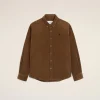 Brown corduroy button-down shirt on hanger against a pale beige background, small dark emblem on the left chest.