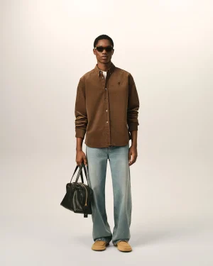 Man standing and facing the camera in a brown corduroy shirt, light jeans, tan shoes, and sunglasses while holding a black bag by his side.