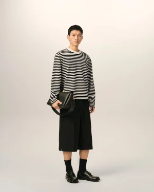 Full-body portrait of a person in a black-and-white striped long-sleeve shirt, black wide-leg pants, black shoes, holding a black bag.