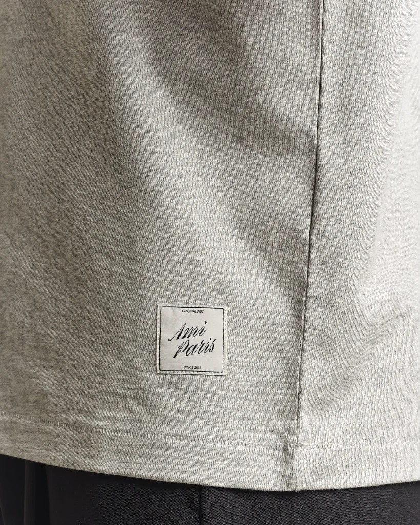 Close-up of heather gray fabric with a stitched seam and a small brand label reading 'Amiri Paris' or 'Amii Paris' on the lower front of the garment.