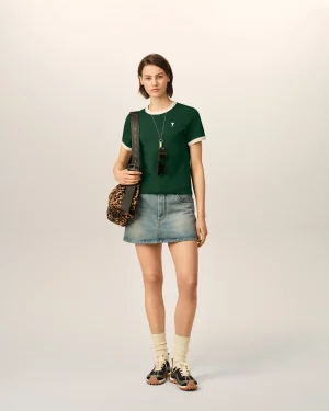 A woman stands against a pale backdrop wearing a green short-sleeve shirt, a denim mini skirt, beige socks, and black sneakers, carrying a leopard-print bag and sunglasses hanging from her neck.