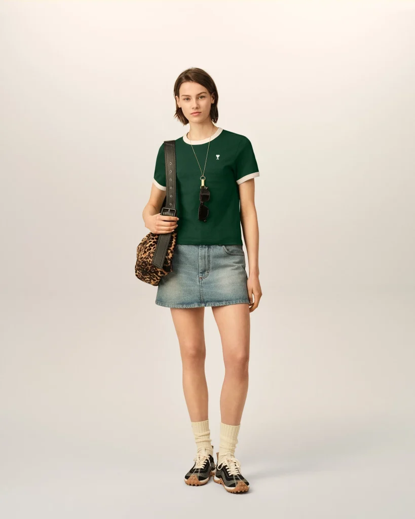 A woman stands against a pale backdrop wearing a green short-sleeve shirt, a denim mini skirt, beige socks, and black sneakers, carrying a leopard-print bag and sunglasses hanging from her neck.