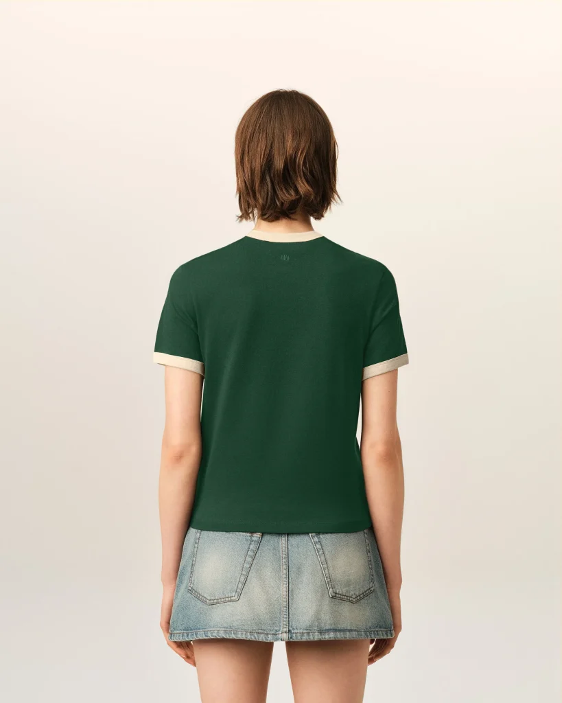 Back view of a person with short brown hair wearing a green T-shirt and a light denim skirt, standing against a pale background.