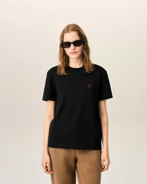 Person wearing a black T-shirt with a small red heart emblem on the chest, sunglasses, and neutral expression against a light background.