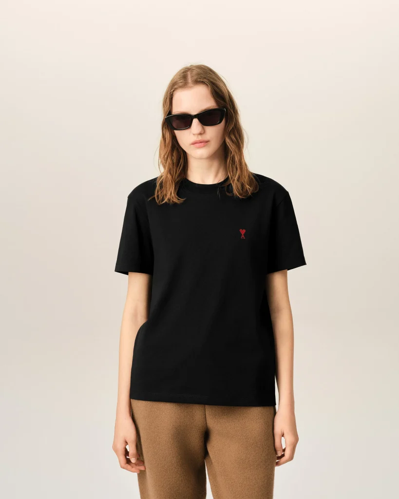 Person wearing a black T-shirt with a small red heart emblem on the chest, sunglasses, and neutral expression against a light background.