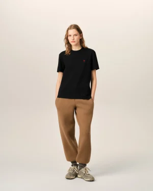 Young woman wearing a black T-shirt with a small red logo, tan/brown pants, and sneakers, standing against a pale beige background.