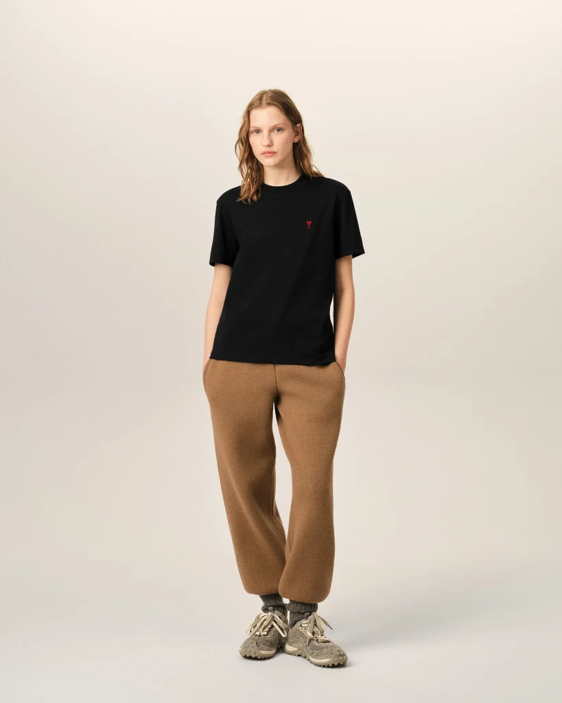 Young woman wearing a black T-shirt with a small red logo, tan/brown pants, and sneakers, standing against a pale beige background.