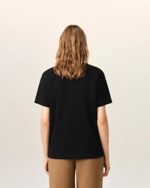 Person viewed from behind wearing a black short-sleeve shirt and light brown pants, against a pale neutral background.