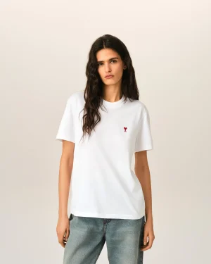 Young woman with long dark hair wearing a white T-shirt featuring a small red heart embroidery on the chest, standing against a beige background.