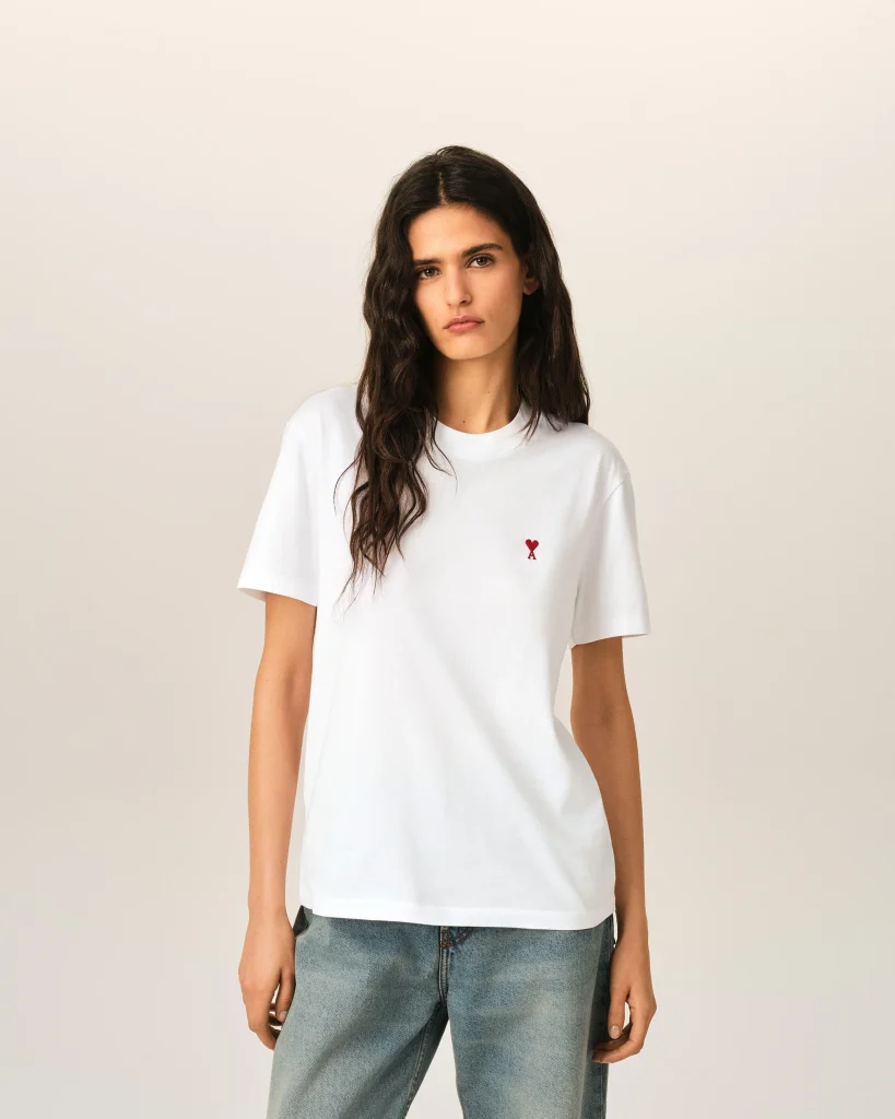 Young woman with long dark hair wearing a white T-shirt featuring a small red heart embroidery on the chest, standing against a beige background.