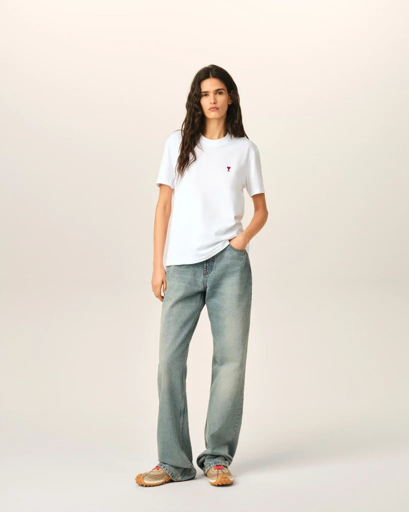Full-body portrait of a woman in a white T-shirt with a small red heart, faded jeans, and tan sneakers against a beige backdrop.