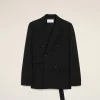 Black double-breasted blazer laid flat on a pale beige backdrop, showing lapels, pockets, and buttons with a white label at the neck area.