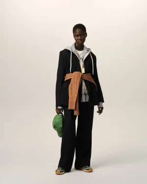 Full-body shot of a dark-skinned person modeling a layered outfit: black blazer over a gray hoodie with a striped shirt, tan knit scarf tied around the waist, green cap in one hand, green plaid sneakers.