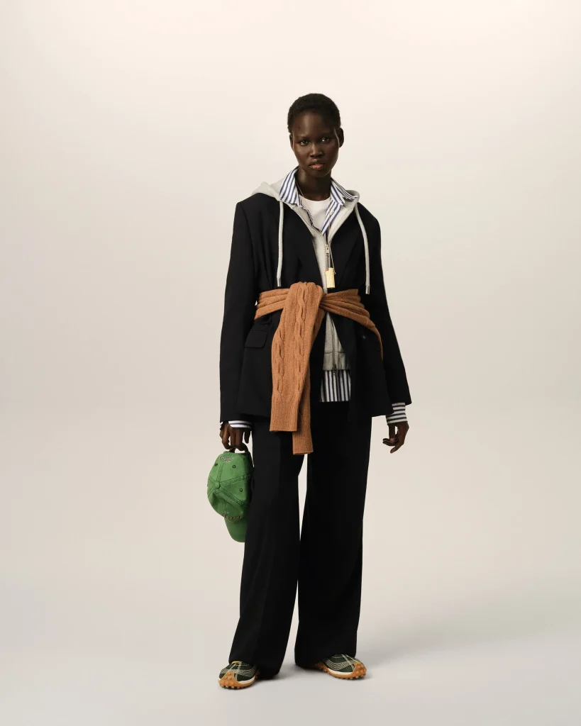 Full-body shot of a dark-skinned person modeling a layered outfit: black blazer over a gray hoodie with a striped shirt, tan knit scarf tied around the waist, green cap in one hand, green plaid sneakers.