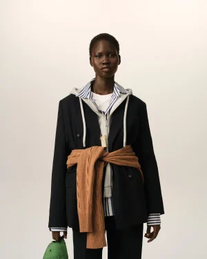 Portrait of a young Black person standing against a light beige background, wearing a striped shirt under a blazer with a gray hoodie and a tan knitted scarf tied around the waist, holding a green hat in the left hand.