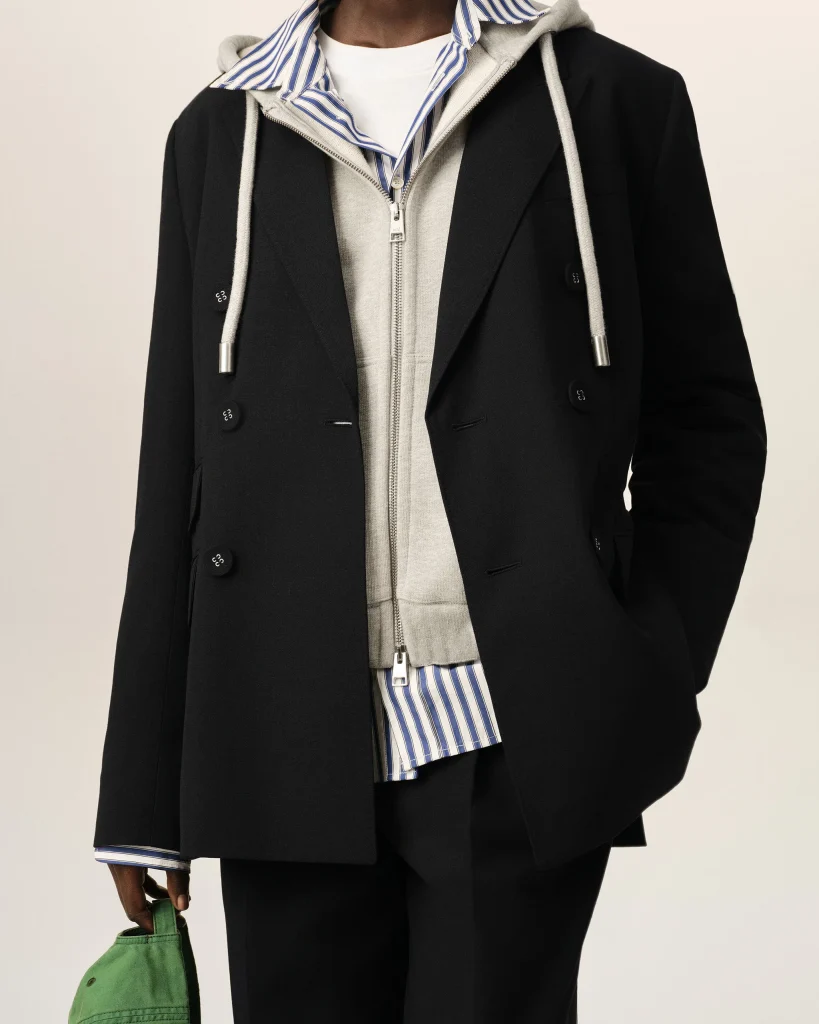 Person wearing a black coat over a gray zip hoodie and blue-striped shirt, holding a green bag at the side.