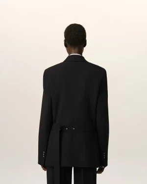 Person seen from behind, wearing a tailored black blazer with a belt against a pale, neutral backdrop.