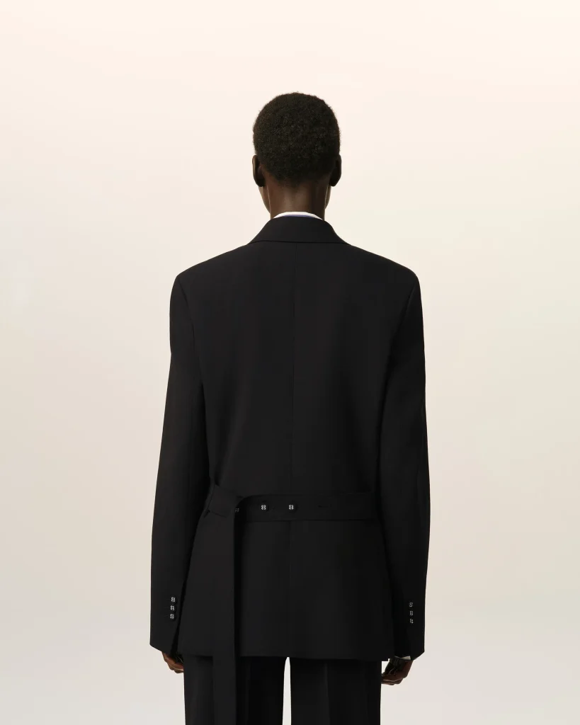 Person seen from behind, wearing a tailored black blazer with a belt against a pale, neutral backdrop.