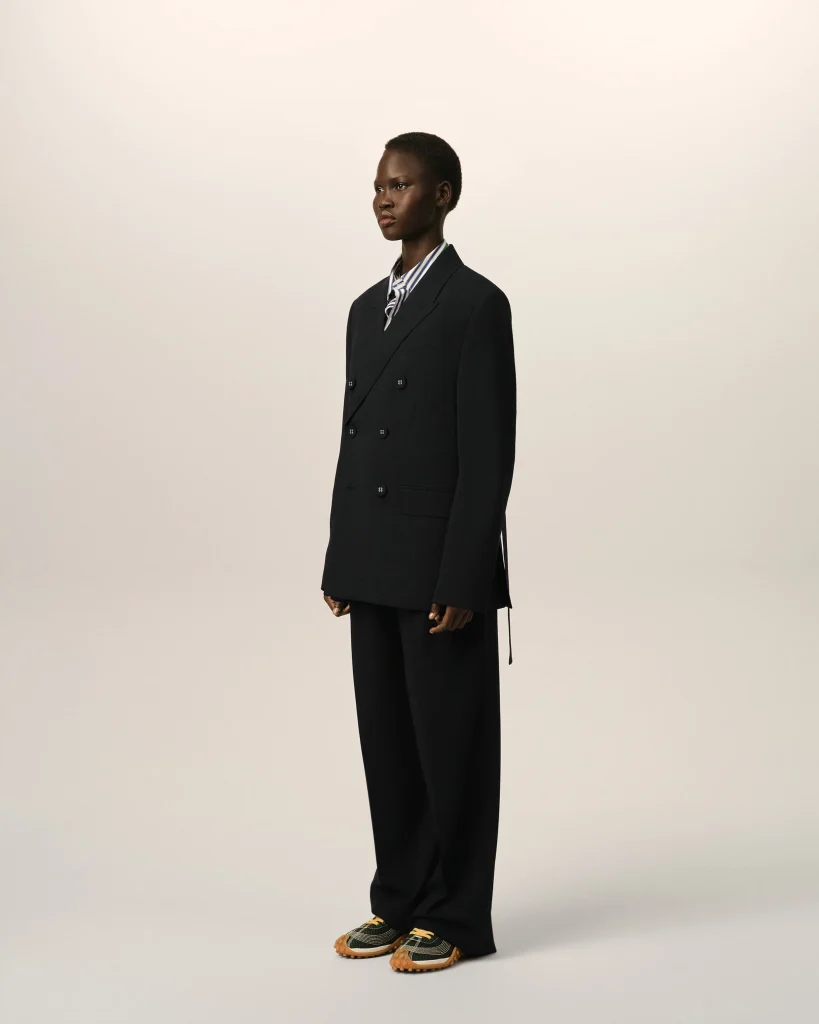 Full-body portrait of a person in a dark oversized double-breasted suit with a striped shirt, standing against a neutral backdrop.