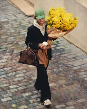 Person walking on a cobblestone street, wearing a green cap and black coat, holding a large bouquet of yellow flowers wrapped in brown paper and a coffee cup.