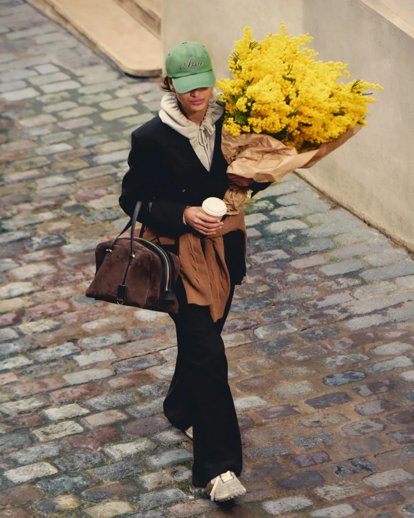 Person walking on a cobblestone street, wearing a green cap and black coat, holding a large bouquet of yellow flowers wrapped in brown paper and a coffee cup.