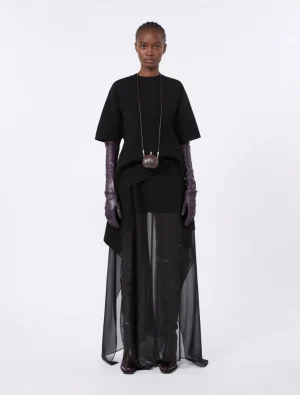 Model wearing a black oversized top, sheer floor-length skirt, purple gloves, and a small brown pendant bag on a long chain.