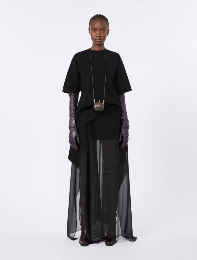 Model wearing a black oversized top, sheer floor-length skirt, purple gloves, and a small brown pendant bag on a long chain.