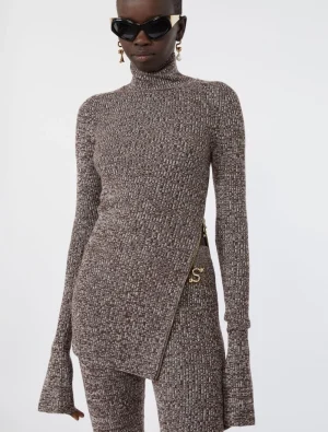 Woman wearing a brown patterned turtleneck knit top with an asymmetric wrap front and matching pants, accessorized with large sunglasses and gold earrings on white background.