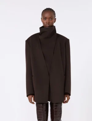 Model wearing a dark brown oversized coat with a high collar, standing against a light background.