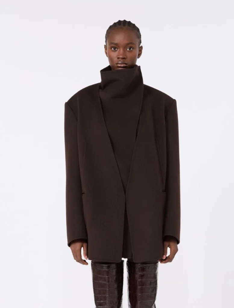 Model wearing a dark brown oversized coat with a high collar, standing against a light background.