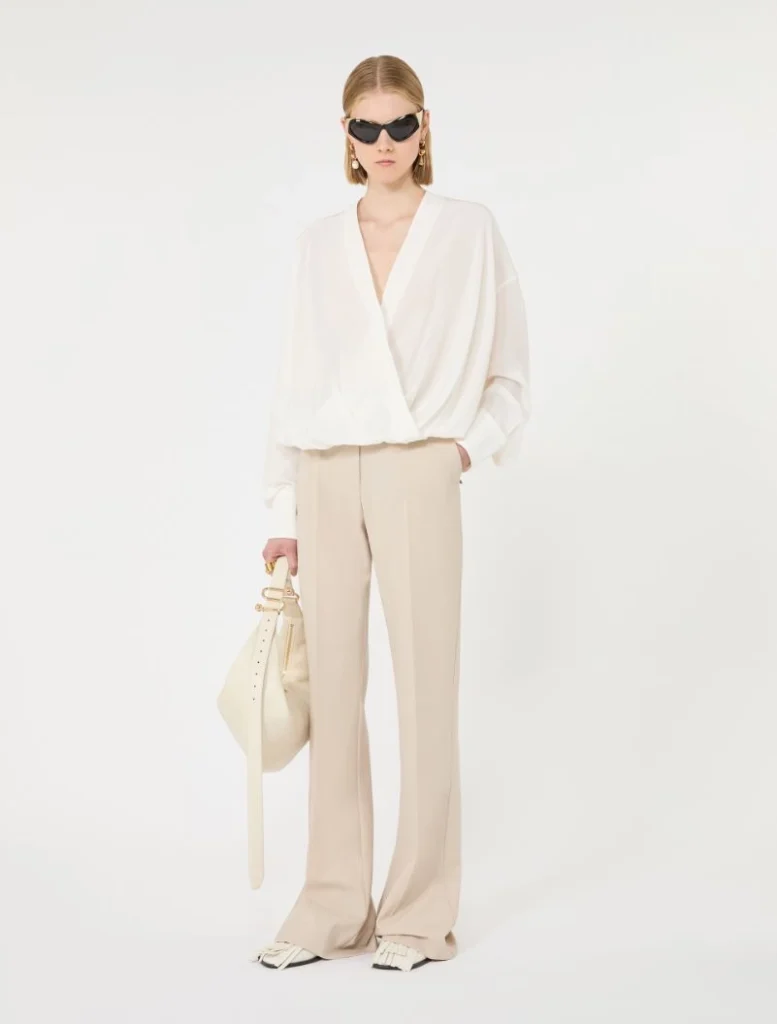 Model wearing a white draped wrap blouse with beige wide-leg trousers and a light beige handbag, posing in sunglasses against a white background.