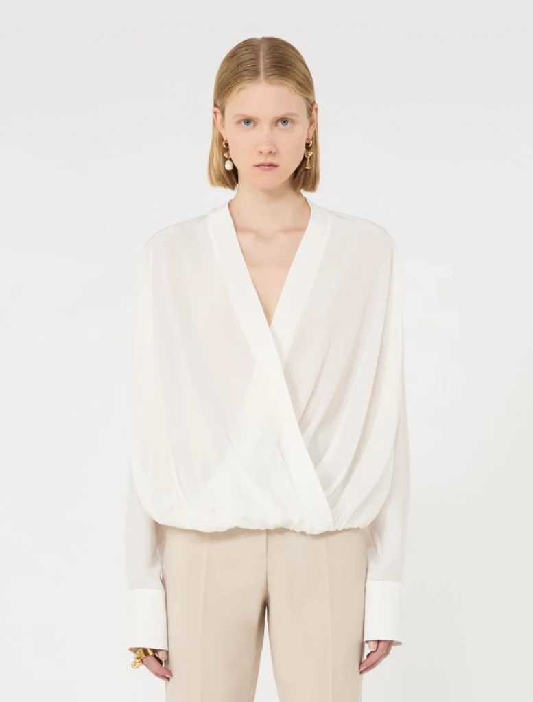Model wearing a white draped blouse with a V-neck, tucked into beige trousers, accessorized with gold earrings and rings.