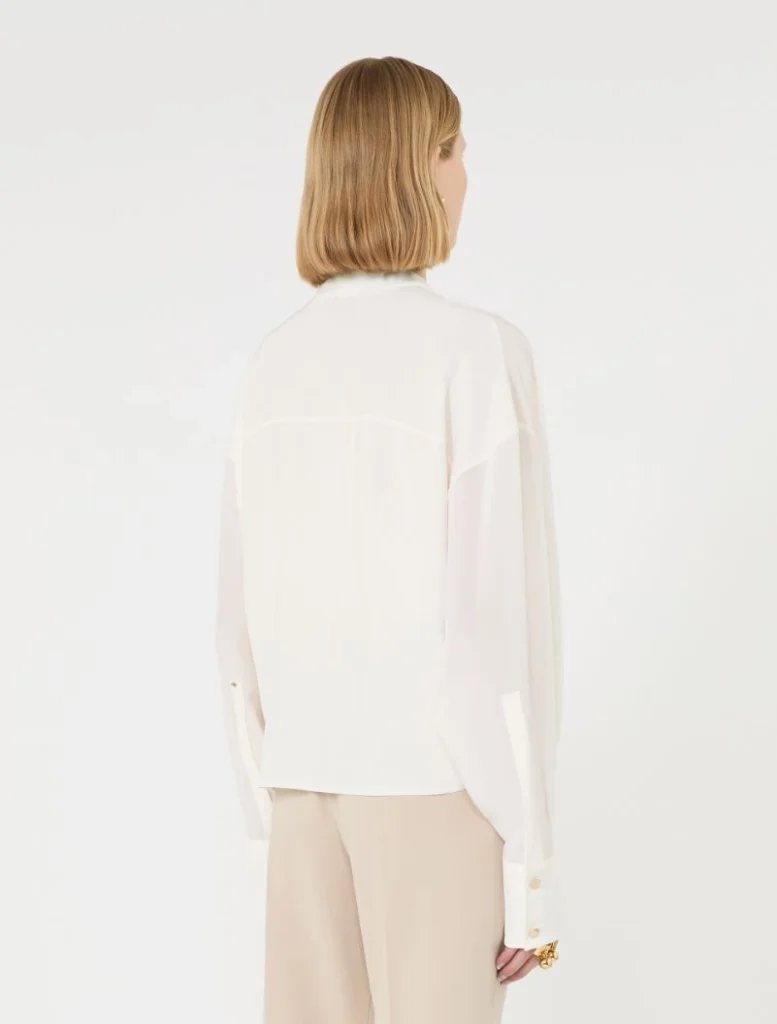 Back view of a person wearing a white blouse and beige wide-leg pants in a studio setting with a white background, blonde hair above the blouse neckline.