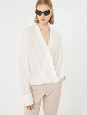 Woman wearing a white wrap blouse with billowy sleeves and beige pants, accessorized with black sunglasses and gold jewelry, standing against a light background.