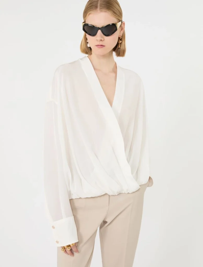 Woman wearing a white wrap blouse with billowy sleeves and beige pants, accessorized with black sunglasses and gold jewelry, standing against a light background.