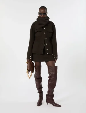 Model wearing a black wool coat with silver buttons, a matching mini skirt, burgundy over-the-knee boots, a large scarf, sunglasses, and a brown clutch with a gold chain in a studio setting.