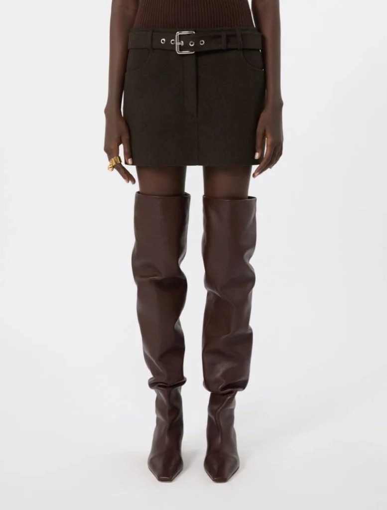 Model wearing a dark mini skirt with a wide belt and dark brown knee-high boots, standing straight.