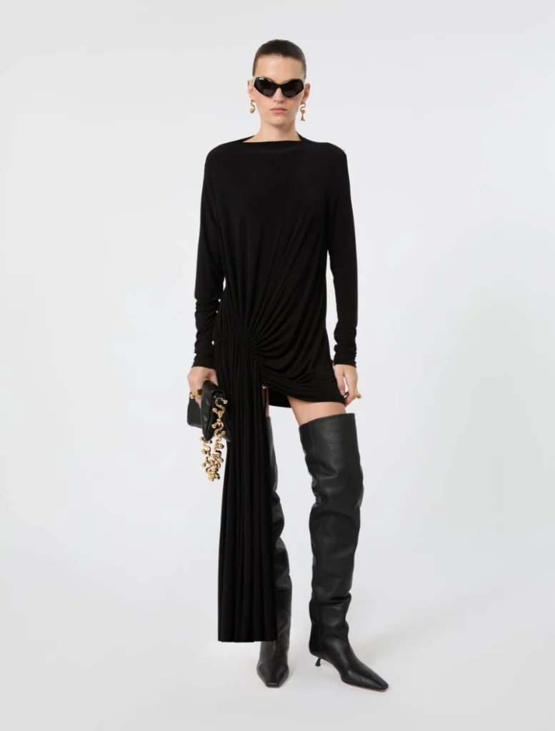 Model in a black draped mini dress with a long front panel, paired with thigh-high black boots and oversized sunglasses.