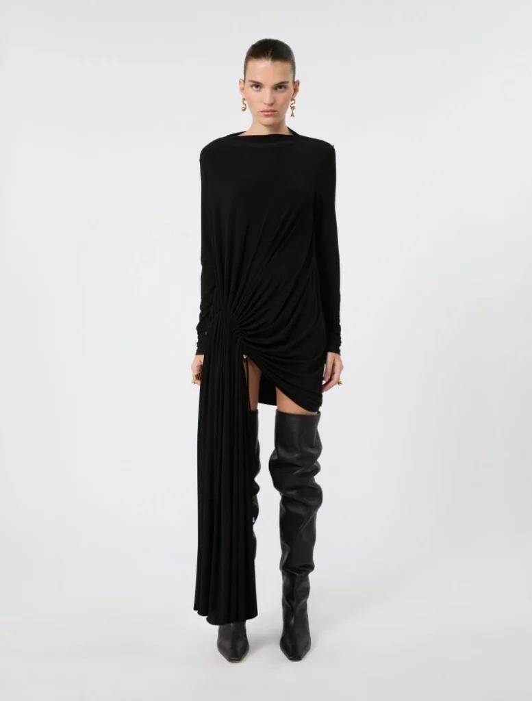 Model wearing a black long-sleeve draped dress with a twisted knot at the hip and a high side slit, paired with black over-the-knee boots and gold earrings.