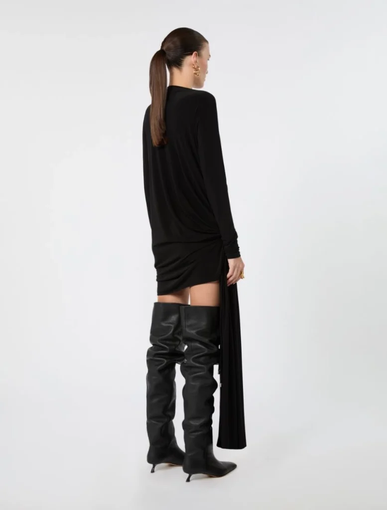 Back view of a model in a black draped long-sleeve mini dress paired with over-the-knee black leather boots, against a light background.