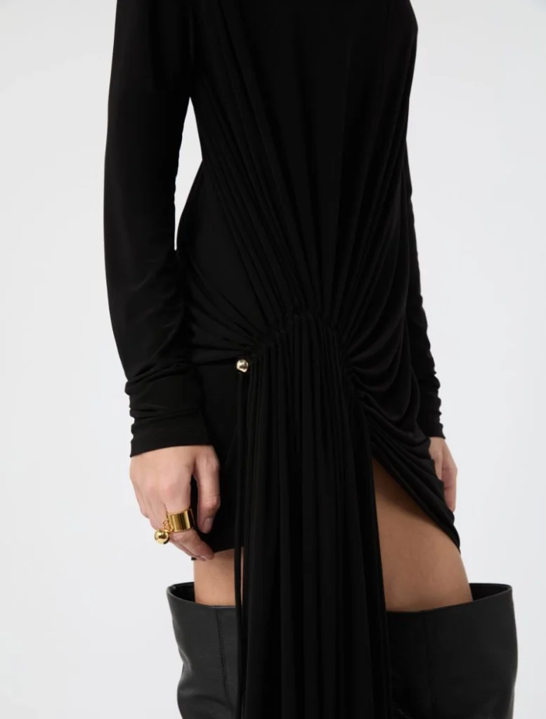 Black draped dress with side ruching and a high slit, shown with a gold ring and black boots nearby.