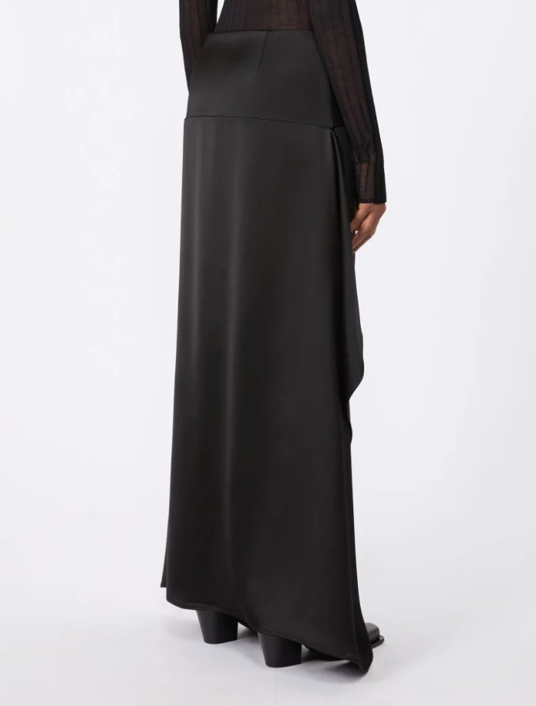 Side view of a person wearing a black satin maxi skirt with a high waist and draped hem, paired with black block-heeled boots.
