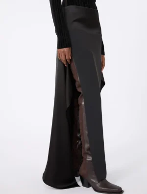 Model wearing a black, long panel skirt with a brown leather panel along the side slit, paired with brown ankle boots