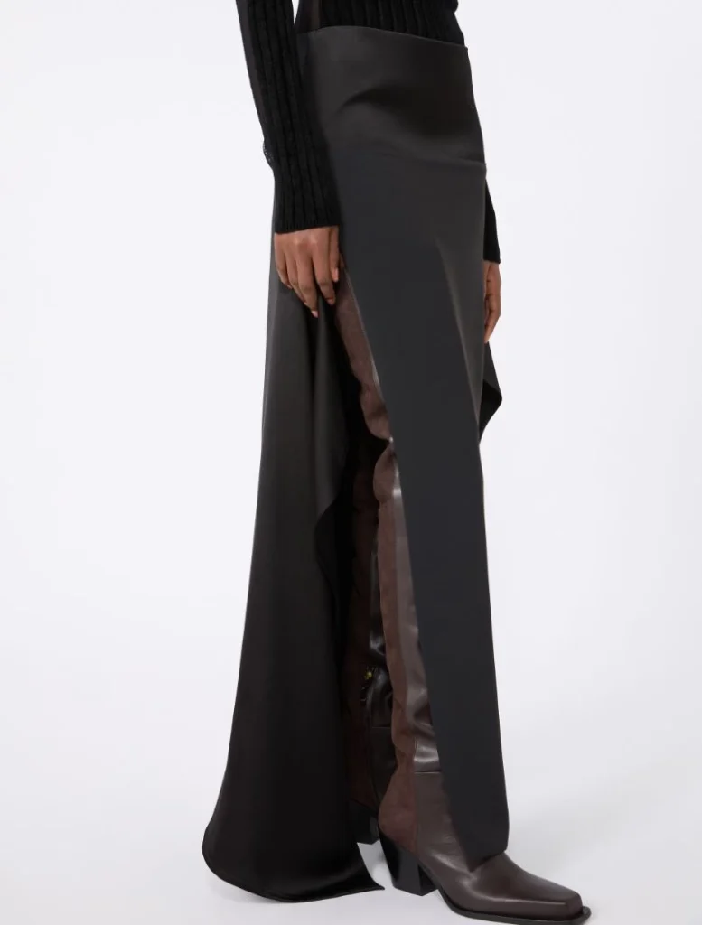 Model wearing a black, long panel skirt with a brown leather panel along the side slit, paired with brown ankle boots