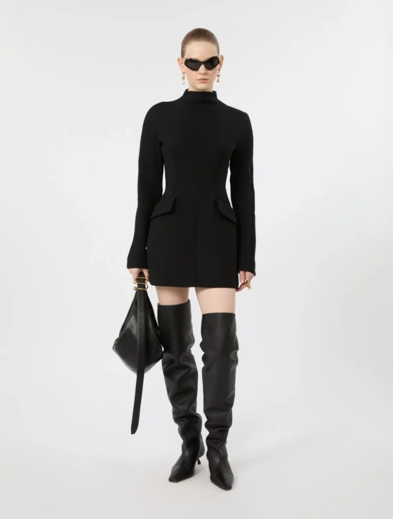 Model wearing a fitted black mini dress with a high neck and structured seams, paired with black over-the-knee boots and a black handbag with gold hardware, sunglasses.