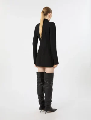 Woman seen from behind wearing a black tailored mini dress and thigh-high black boots against a white studio backdrop.