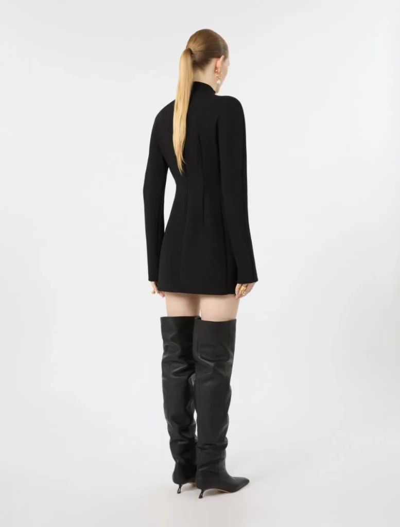 Woman seen from behind wearing a black tailored mini dress and thigh-high black boots against a white studio backdrop.
