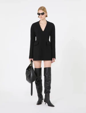 Model wearing a black V-neck blazer dress with long sleeves, knee-length, paired with tall black boots and oversized sunglasses, carrying a black handbag.