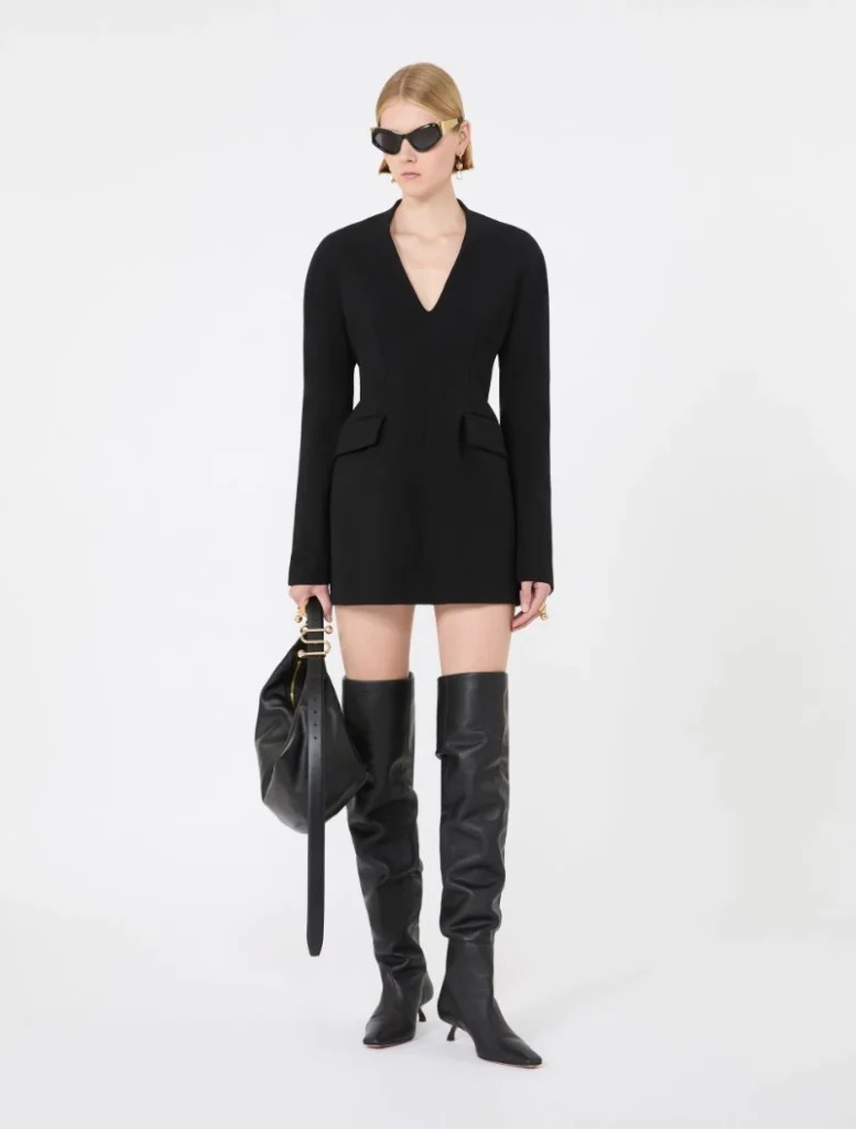 Model wearing a black V-neck blazer dress with long sleeves, knee-length, paired with tall black boots and oversized sunglasses, carrying a black handbag.