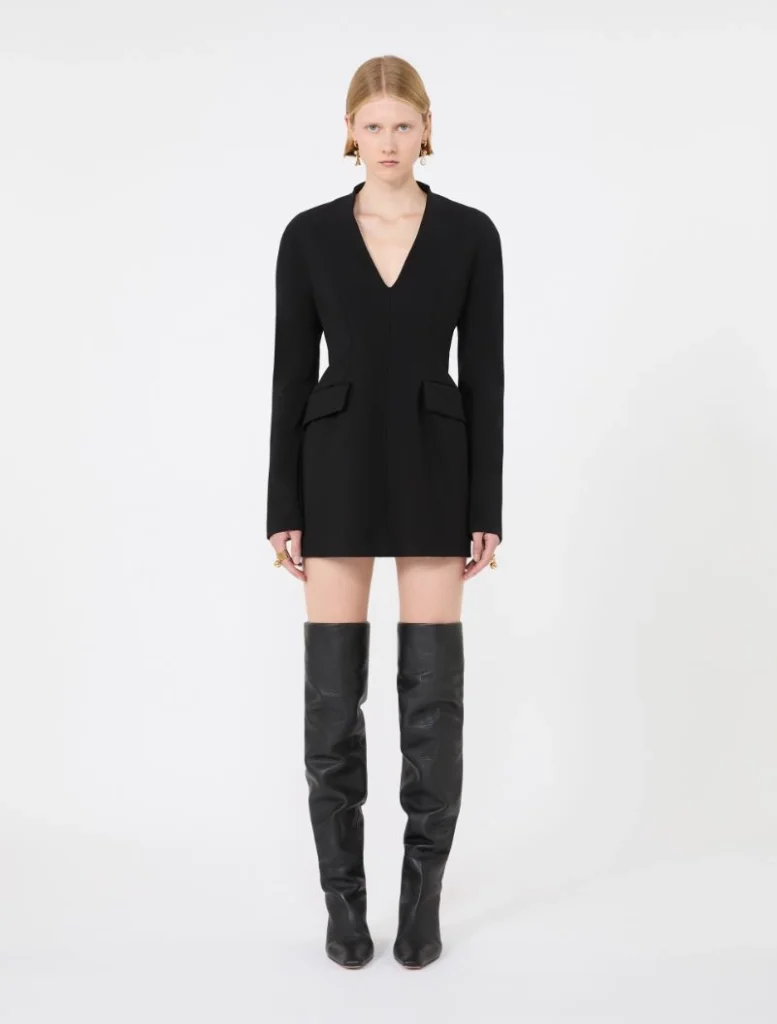 Model in a black tailored mini dress with long sleeves, deep V-neck, and front pockets with flaps, paired with black thigh-high boots, standing against a white studio background.